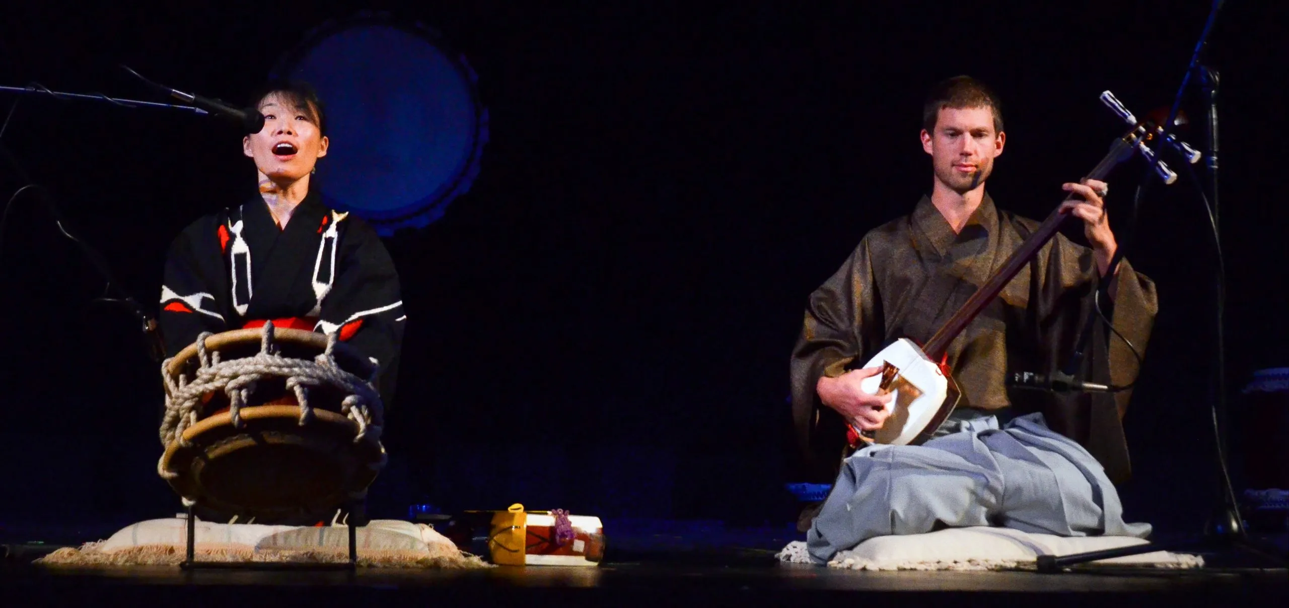 Kyle performing shamisen on a dark stage beside a taiko drummer